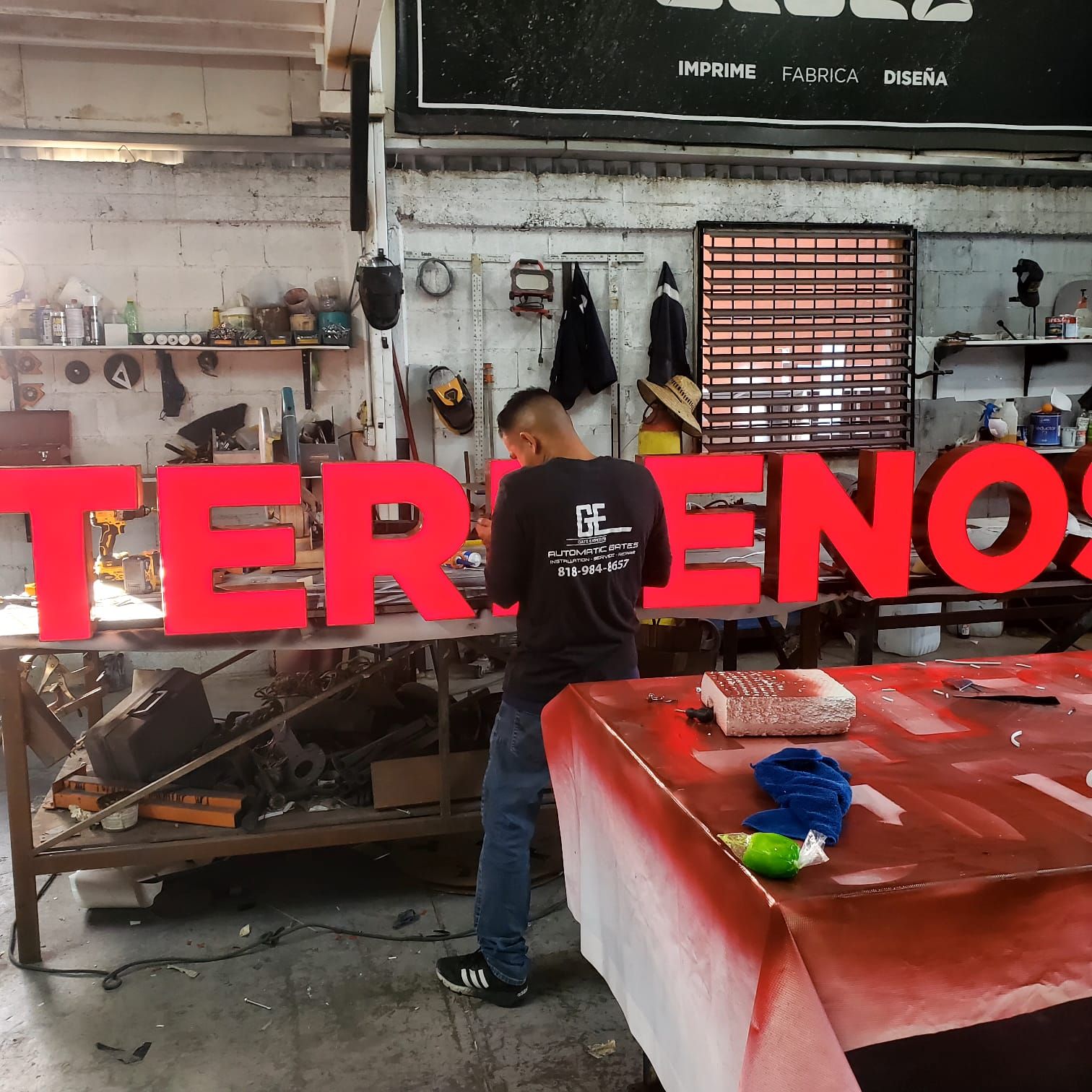 Armado en banco de las letras de canal rojas de "TERRENOS", encendidas para prueba mientras el técnico sella una pieza.