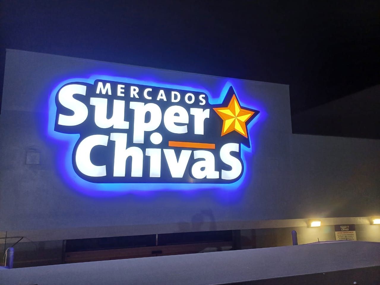 Anuncio de Mercados Super Chivas de noche: letras blancas con retroiluminación azul y estrella amarilla, sobre muro liso.